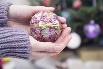 Decorating a Christmas tree at home.Hands holding a vintage pink Christmas-tree ball with glitter and a flower pattern. A blurred fir-tree with balls in the back. Close up