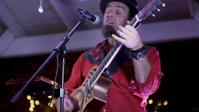 Closeup of guitarist playing resonator and singing at an outdoor concert at a restaurant at night.