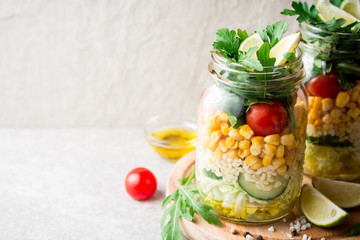 Healthy homemade salads with bulgur, vegetables and lime in mason jars on grey stone background.