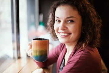 Beautiful girl by the window in a cafe holds a big cup and smiles.