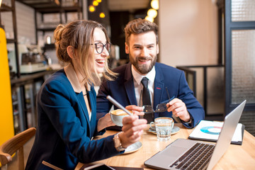 Caucasian business couple dressed strictly in the suits working together with laptop and paper charts sitting at the cafe during the coffee break