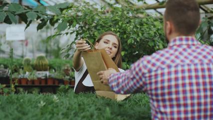 Obraz premium Young smiling seller florist working in garden center. Woman gives shopping bag to customer at store