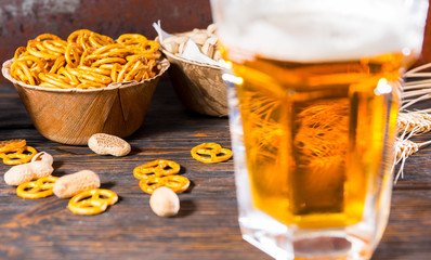 Focus on plate with small pretzel near plates with pistachios and glass with light beer and a head of foam on dark wooden desk