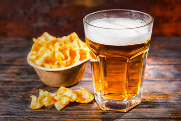 Big glass with freshly poured light beer near plate with chips on dark wooden desk
