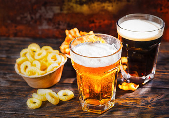 Two glasses with freshly poured light and dark beer near plates with snack and chips on dark wooden desk