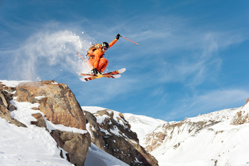 A professional skier makes a jump-drop from a high cliff against a blue sky leaving a trail of snow...