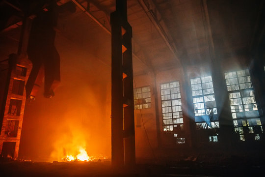 Fire In Large Hall Of Factory Or Warehouse At Night. Huge Flame Of Red And Yellow Fire And Blue Light From Windows