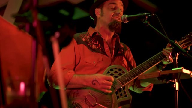 Looking through the female double bass, a male roots musician rocks out on a resonator guitar at night at a restaurant.