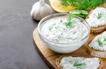 Obraz na płótnie Canvas Homemade greek tzatziki sauce in a glass bowl with ingredients and sliced bread on a dark black stone background. Cucumber, lemon, dill, garlic. Close-up, horizontal, selective focus on a bowl
