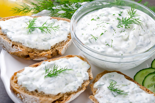 Homemade greek tzatziki sauce in a glass bowl with ingredients and sliced bread on a dark black stone background. Cucumber, lemon, dill, garlic. Close-up, horizontal, selective focus on a bowl