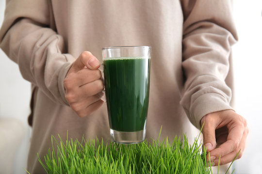 Woman holding glass cup of fresh juice over sprouted wheat grass, close up