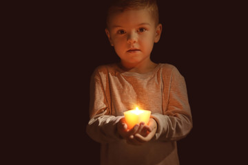 Little boy holding burning candle in darkness