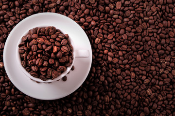 White coffee cup and saucer one single for espresso or capuccino filled with roasted beans on a background of scattered dark coffee beans photo top view