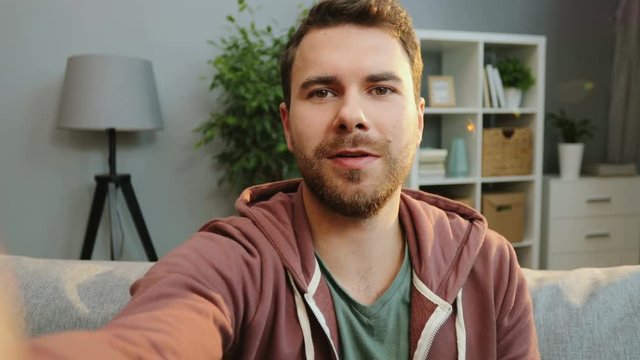 Close up of the young caucasian man having video chat and pointing at something while siting on the sofa in the cozy living room. PVO. Portrait. Indoors