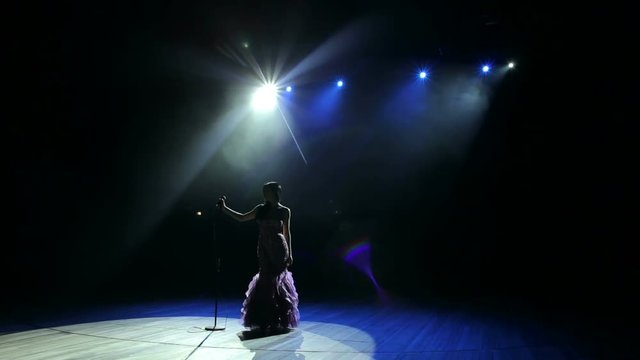 Luxurious singer stands on stage in a long dress in the dark and holding a microphone stand. Wide shot.