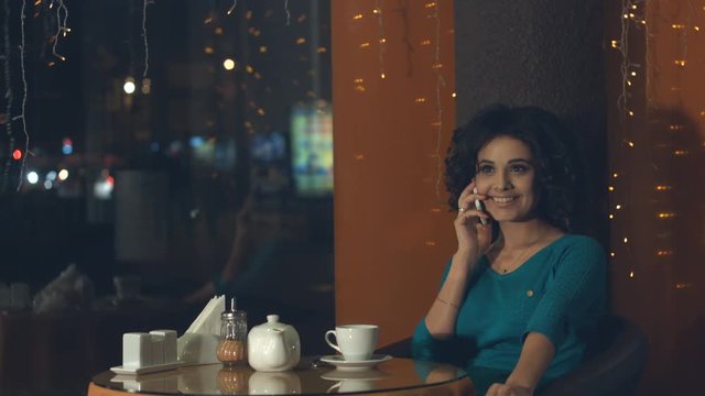 Girl with a smartphone in a cafe.
A pretty girl is sitting in a cafe and talking on the phone. She smiles and drinks tea.
Outside the window lights the evening city.