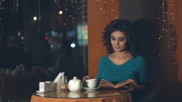 Girl with a book in a cafe.
Slow motion. A pretty girl is sitting in a cafe and reading a book.
Outside the window lights the evening city.