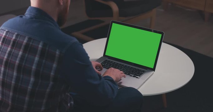 Over the shoulder shot of a young Caucasian man sitting on sofa, working from home on a laptop, green screen chroma key. 4K UHD 60 FPS SLO MO