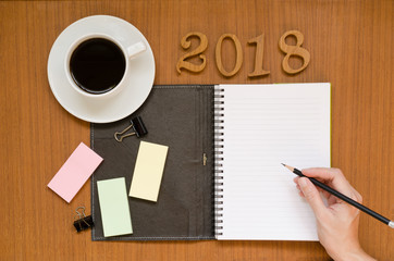Recycled notebook with woman hand writing in the empty paper note surrounding with black coffee in white cup, 2018 letters, yellow, pink, green sticky pad, pencil & paper clip on wood background space