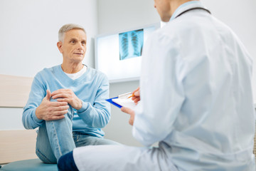 Obraz premium Pain in the knee. Good looking nice aged man holding his knee and looking at his doctor while explaining where he feels pain