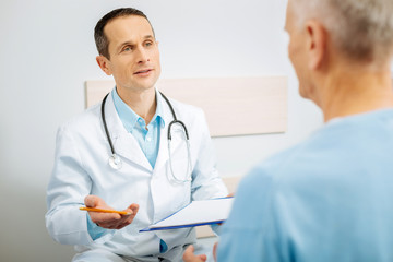 Professional treatment. Nice handsome smart doctor sitting opposite his patient and talking to him while holding his notes