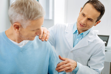 Obraz premium Do you feel anything. Cheerful positive handsome doctor holding patients arm and checking it while standing behind his elderly patient