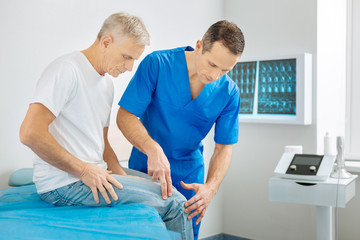 Fototapeta premium Absolutely concentrated. Serious pleasant male therapist standing near his patient and checking his leg while focusing on his work