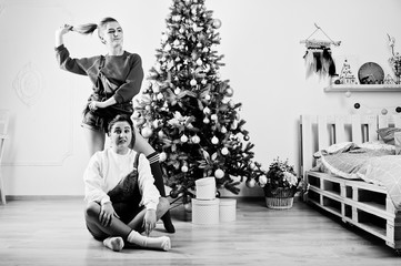 Two fun beautiful girls friends wear in overalls jeans shorts and gaiters against new year tree with chrisrmas decoration.