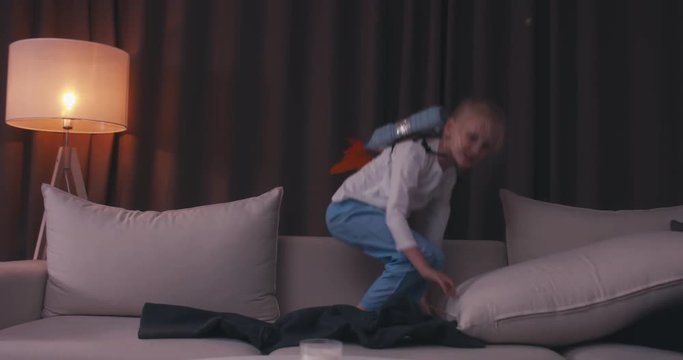 Funny cute dreamer girl wearing rocket pack made from plastic bottles pretending to be an astronaut or pilot, jumping from sofa. 4K UHD 60 FPS SLO MO