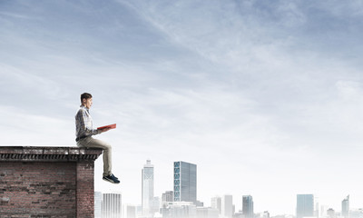 Man on roof edge reading book and cityscape at background