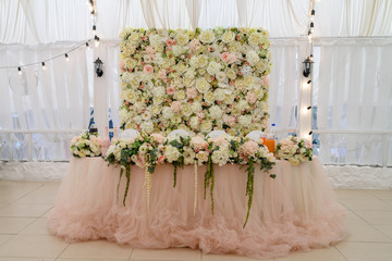 Wedding presidium in restaurant, free space. Wedding banquet table for newlyweds with flowers, greenery, pink cloth and bulbs. Lush floral arrangement on wedding table. Luxury wedding decorations