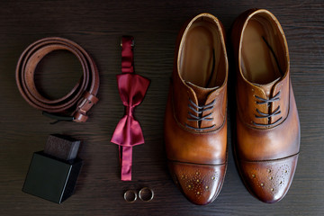 Close up of groom brown leather shoes, bowtie, belt, parfume and golden wedding rings. Modern man or groom accessories. Wedding details. Male casual outfits. Top view, flat lay