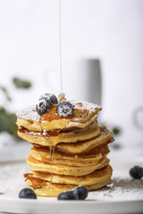 Pile of pancakes with blueberries on the top, maple leaf syrup is pouring on it.