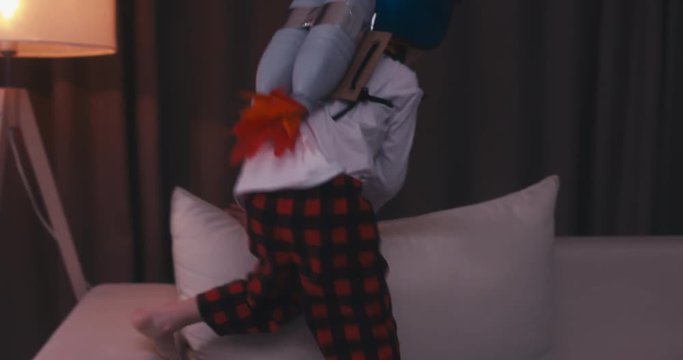 Funny cute dreamer kid boy wearing rocket pack made from plastic bottles pretending to be an astronaut or pilot, jumping from sofa. 4K UHD 60 FPS SLO MO