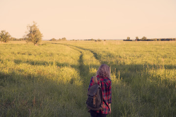 girl in plaid shirt with a backpack on shoulders walking down country road in the middle of a field with tall grass