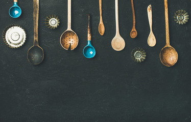 Flat-lay of various old vintage kitchen spoons and baking tin molds over black stone background, top view, copy space. Rustic cooking concept