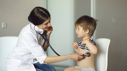 Young woman doctor listening little boy with stethoscope in medical office