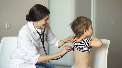 Young woman doctor talking little boy and listening with stethoscope in medical office