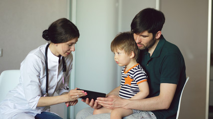 Fototapeta premium Young woman doctor talking with father of little boy using tablet computer in medical office
