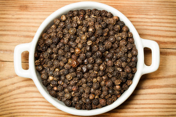 Pimienta negra en grano dentro de un cuenco blanco sobre una tabla de madera. Vista superior