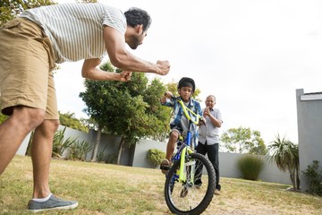 Father and grandfather cheering for boy cycling in yard