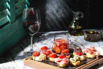Tapas or bruschetta variety. Bread with ham prosciutto, sun dried tomatoes, olive oil, olives, pepper on slate wood board with glass of red wine over white table. Light via old jalousie. Rustic style.