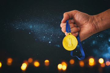 Man hand holding gold medal against black background with glitter overlay. Award and victory concept.