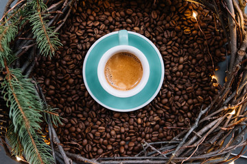 Delicious freshly brewed espresso in a ceramic green cup with saucer, with a beautiful crema, set on coffee beans and with winter festive handmade wooden wreath with fireflies around it, top view