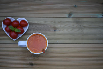 food photography image of homemade tomato soup in a cup with white love heart shape dish filled with fresh vegetable and basil leaves on rustic wood background with space