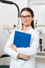 portrait of smiling optometrist in white coat with notepad in hands looking at camera in clinic