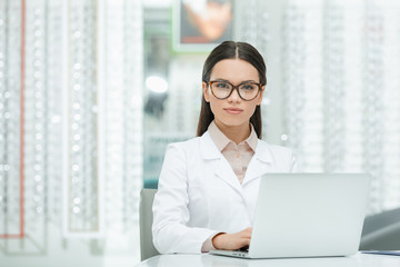 portrait of young ophthalmologist in white coat sitting at table with laptop in optics