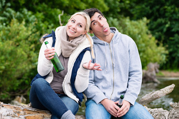 Happy lovers in vacation sitting at waterside on a trunk clinking beer bottles