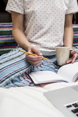 A woman is sitting in bed on a blanket in her hands holding a cup of hot drink and preparing to make a note in a notebook with a pencil, next to her laptop.