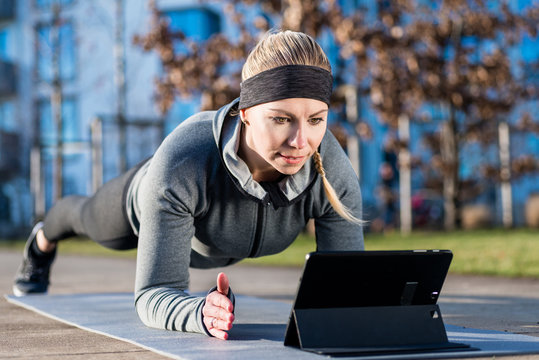 High-angle view of a fit young woman, watching a motivational video on tablet PC while exercising the forearm plank position on a mat outdoors in the park